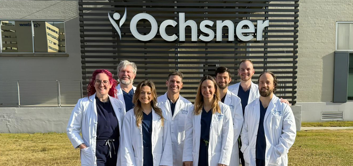 Group of eight Ochsner medical professionals in white lab coats smiling outside the Ochsner Academics building.