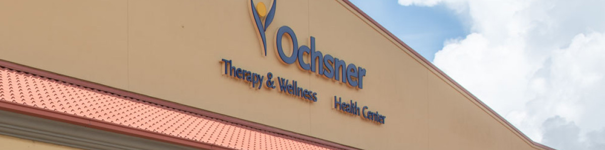 Close-up exterior view of the Ochsner Therapy & Wellness Health Center sign mounted on a beige building facade above a red-tiled roof edge.