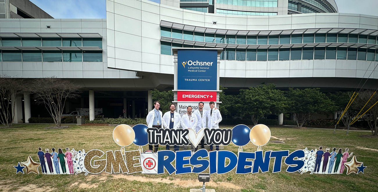 Ochsner Lafayette General Medical Center general surgery residents standing behind a 'Thank You GME Residents' lawn display outside the hospital building.