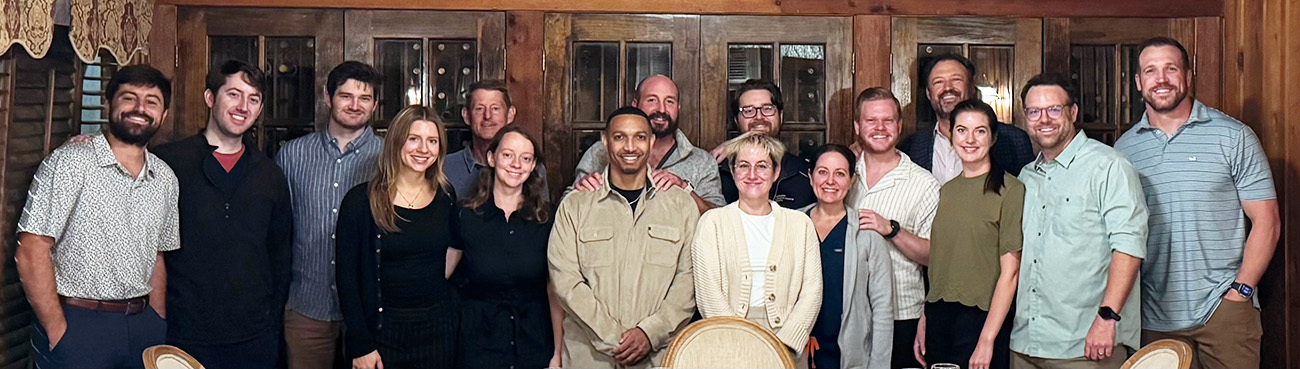A group photograph of General Surgery residents and faculty smiling and posing indoors. The team members are arranged in two rows against a background of warm, wood-paneled walls and traditional cabinets.