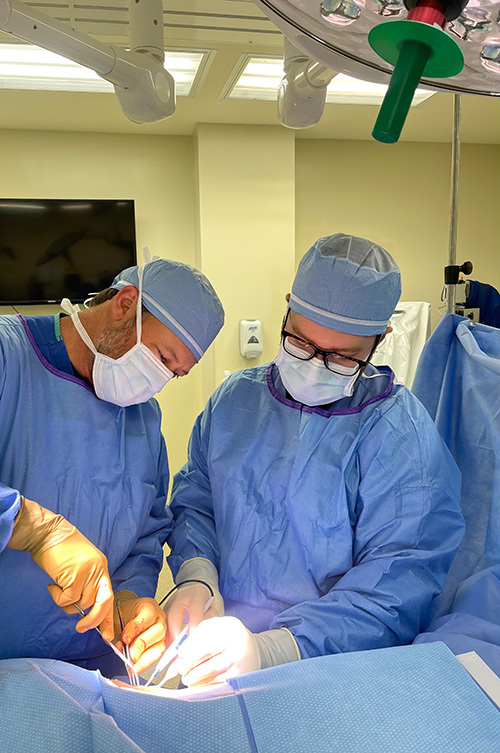 Two surgeons wearing blue scrubs, surgical caps, and face masks, with one wearing glasses, are focused on performing a medical procedure on a patient in a sterile operating room
