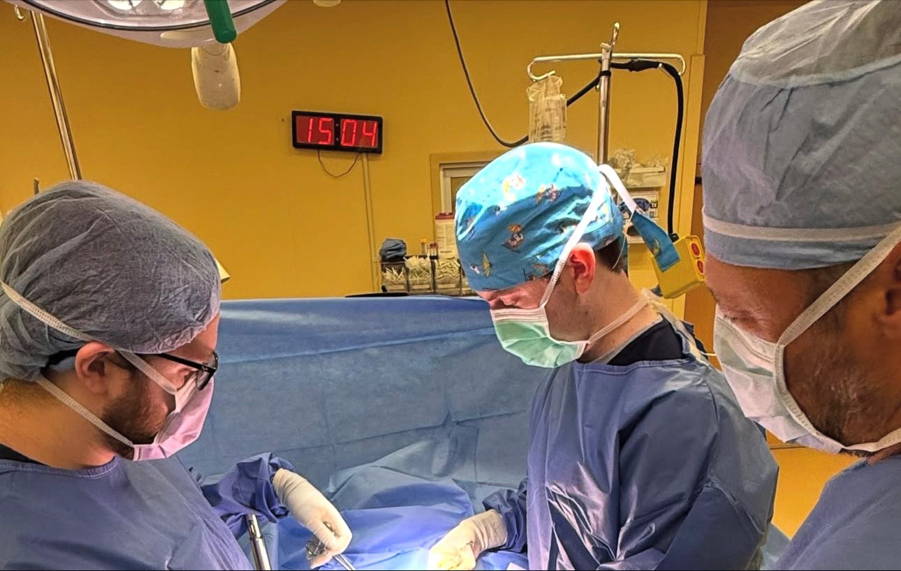Medical students wearing scrubs, caps, and masks performing a medical operation in a brightly lit operating room with a digital clock on the wall.
