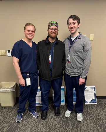 Three male medical professionals smiling and posing together indoors, wearing scrubs and casual hospital attire.