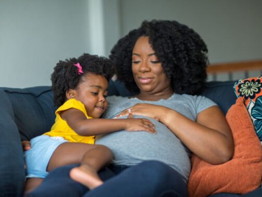 Pregnant mom lying on the couch with her daughter touching her belly.