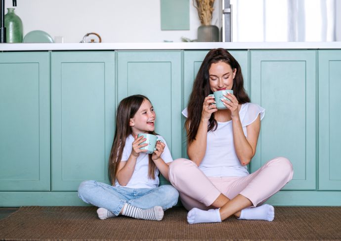 Mother and daughter sitting on the ground in the kitchen drinking coffee together