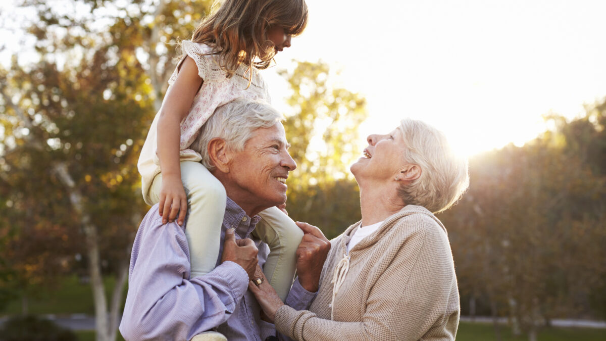 Grandparents outside with their granddaughter, smiling at each other while the granddaughter is on the grandfathers shoulders