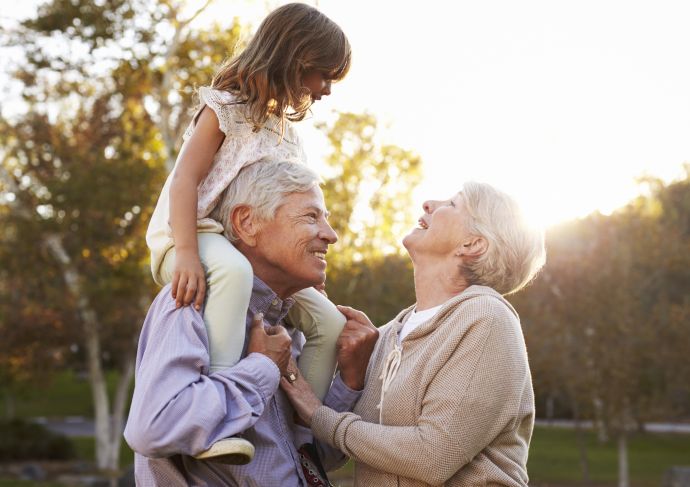 Grandparents outside with their granddaughter, smiling at each other while the granddaughter is on the grandfathers shoulders
