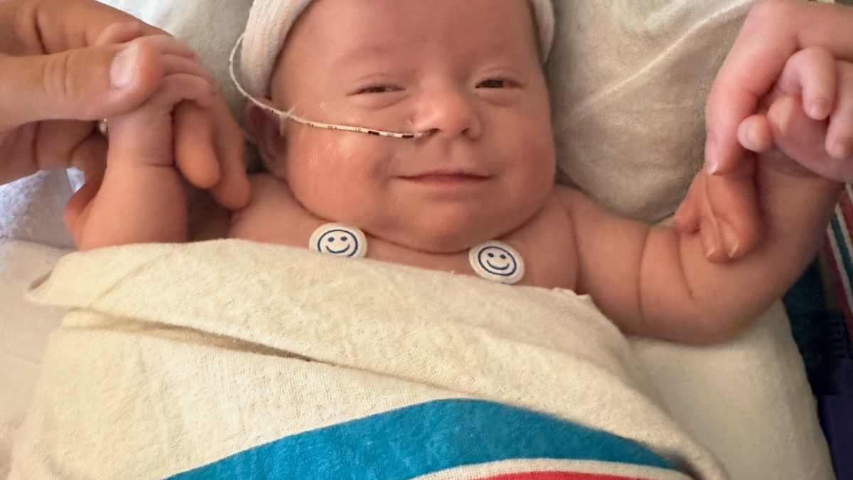 Baby in a hospital bed is holding mom and dad's hands and smiling