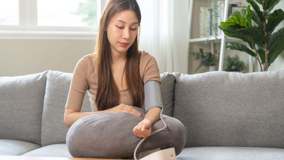 young woman sitting on couch checking blood high pressure and heart rate with digital pressure monitor machine