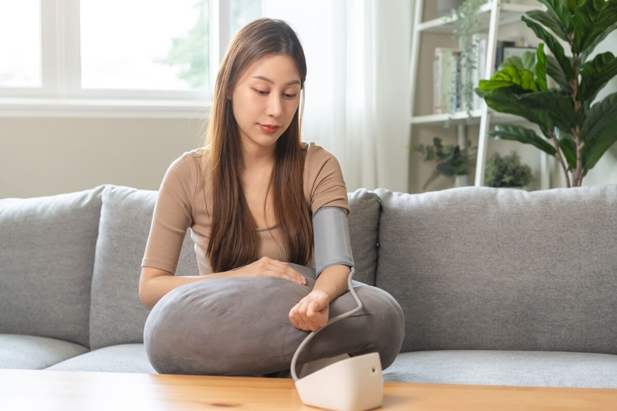 young woman sitting on couch checking blood high pressure and heart rate with digital pressure monitor machine