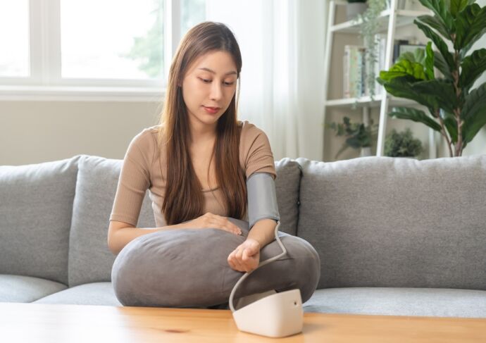 young woman sitting on couch checking blood high pressure and heart rate with digital pressure monitor machine