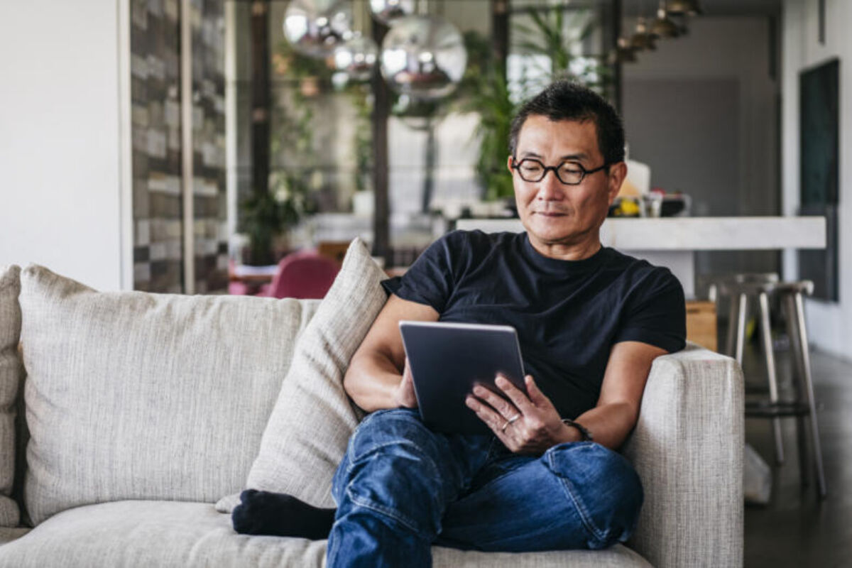 Man sitting on a couch in his house looking at a tablet with glasses on.