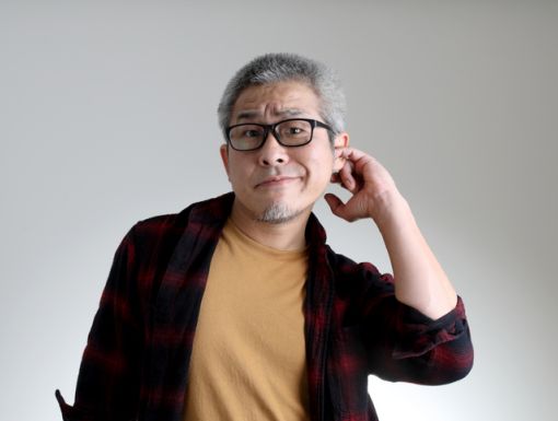 Man with glasses and gray hair scratches ear with his finger