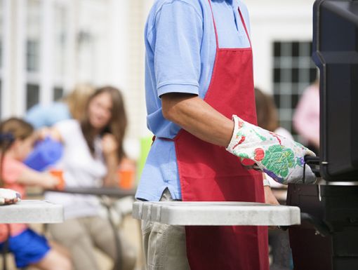 Man grilling wearing flame-retardant mitts and apron with people far away in the background.