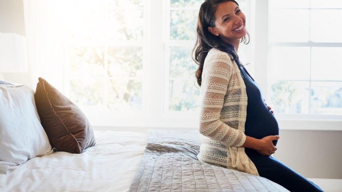 Pregnant woman sitting on the edge of a bed smiling and holding her stomach.