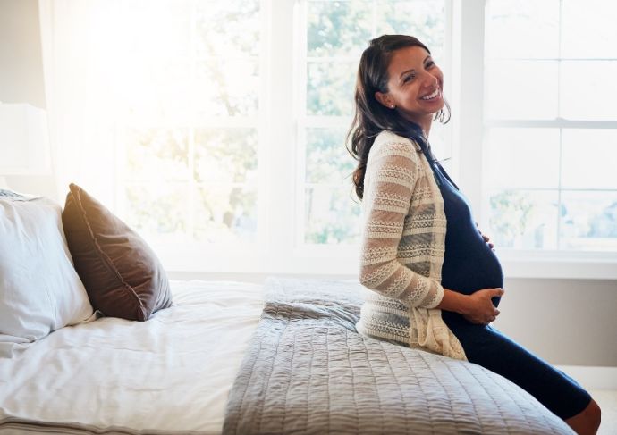 Pregnant woman sitting on the edge of a bed smiling and holding her stomach.