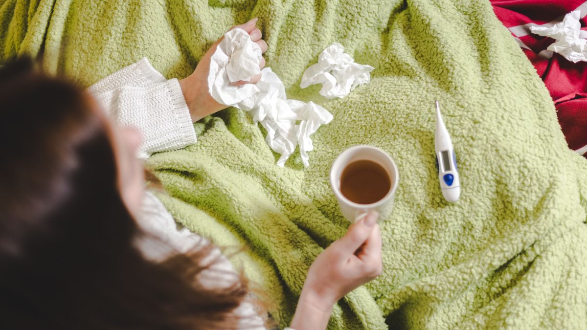 Woman holding a mug and tissue paper in bed with a thermometer next to her