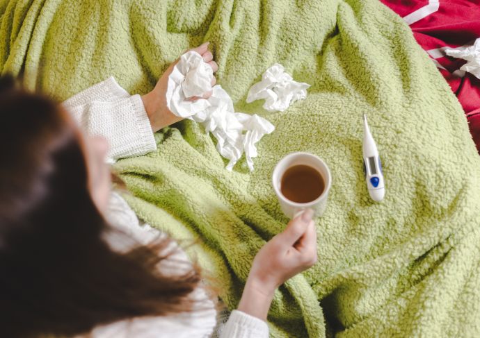 Woman holding a mug and tissue paper in bed with a thermometer next to her