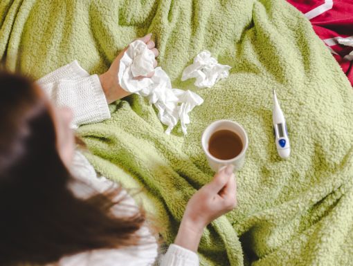 Woman holding a mug and tissue paper in bed with a thermometer next to her