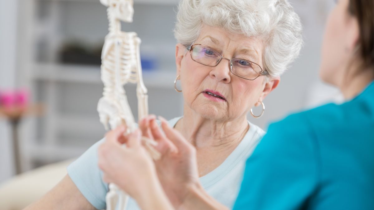 A female provider showing an elderly woman a skeleton model to showcase what happens to bones when you have osteoporosis