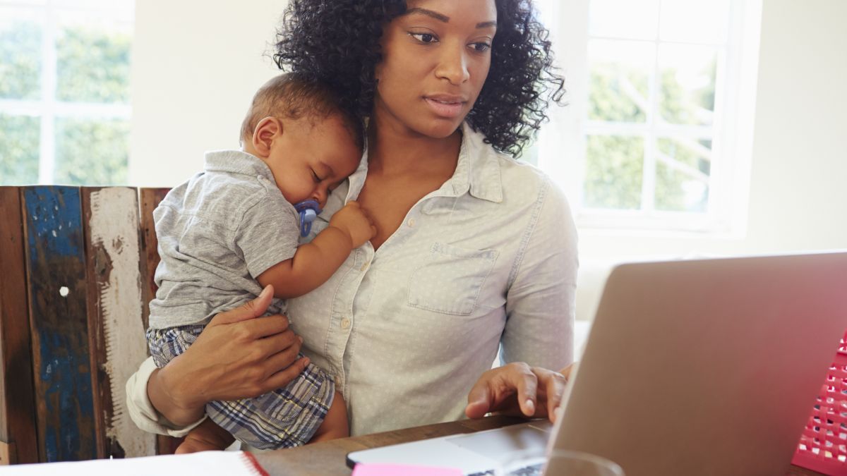 A working mom holding her baby at her desk while looking at her work computer