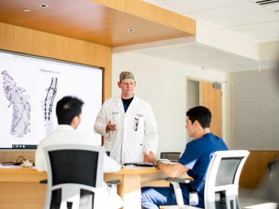 Doctor standing in front of board speaking to two people in chairs