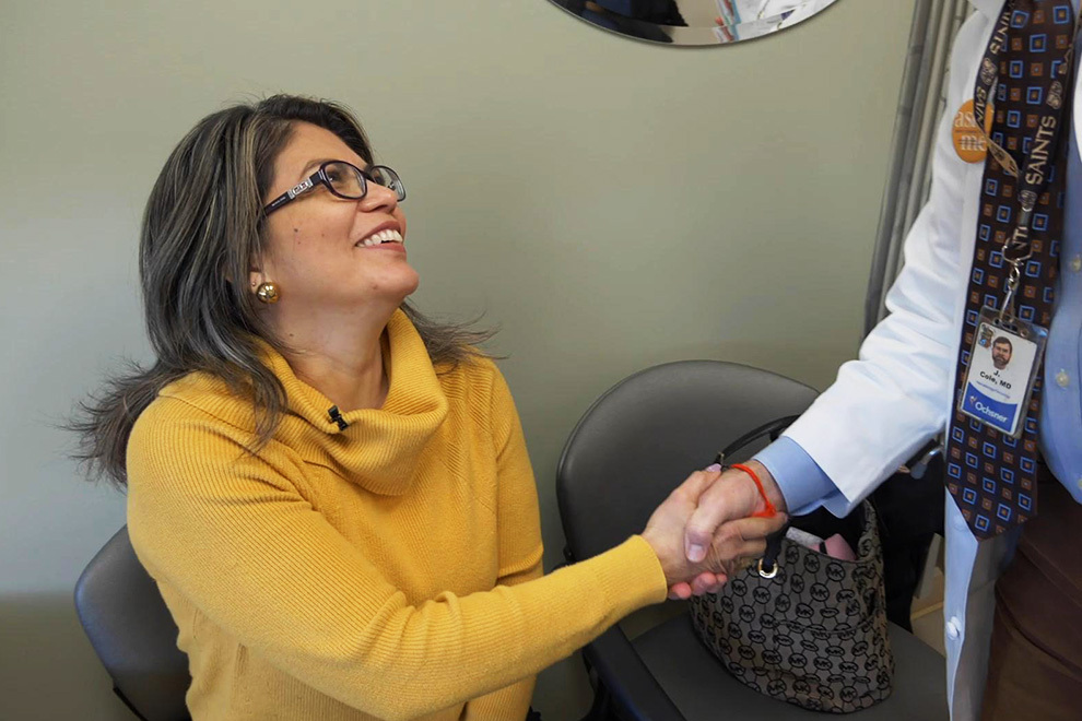 A smiling female patient wearing glasses and a yellow turtleneck sweater seated in a chair, shaking hands with a doctor wearing a white lab coat and an Ochsner ID badge.