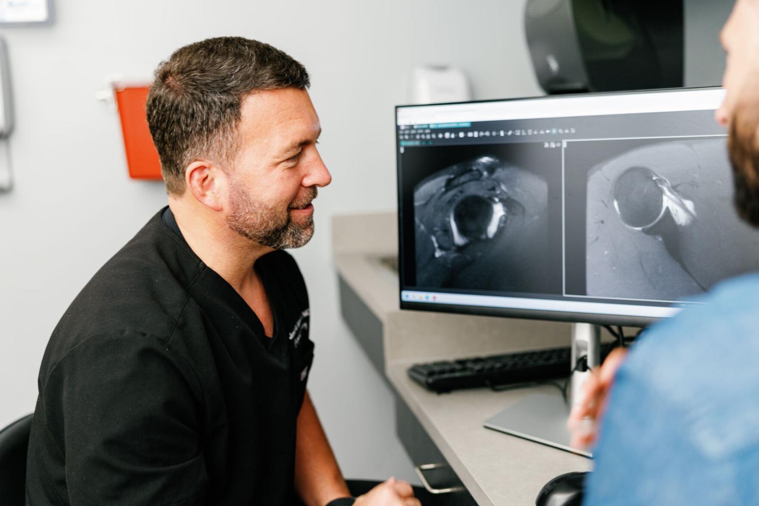 Dr. Jeremy Burnham wearing black scrubs reviewing medical imaging scans on a computer monitor while talking with a patient.