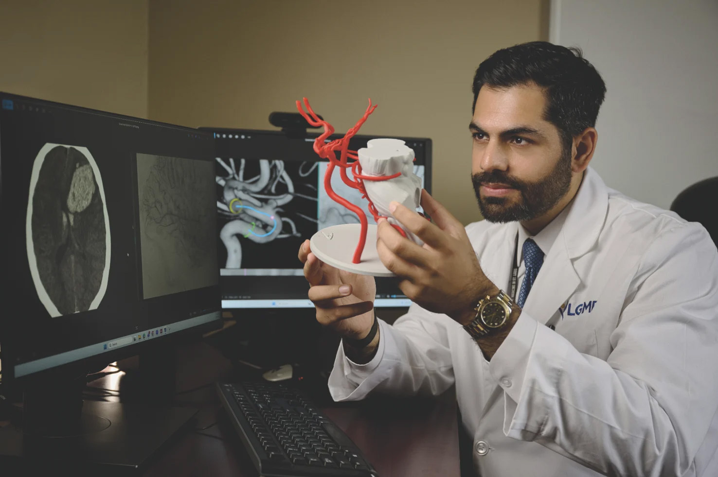 Mohammad AlMajali, MD in a white lab coat sitting at a desk with dual computer monitors displaying brain scans while examining a 3D anatomical model of blood vessels.
