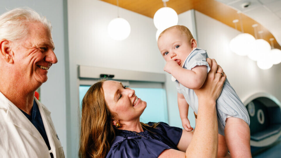 mother holding infant son into the air next to smiling surgeon in a white coat