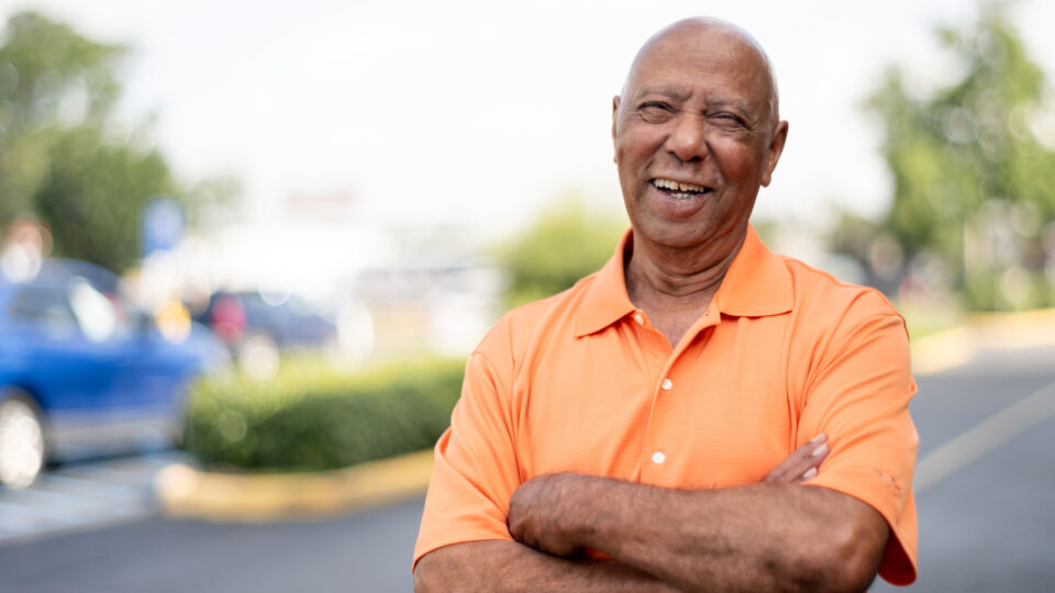Older man in orange shirt standing outside with arms crossed